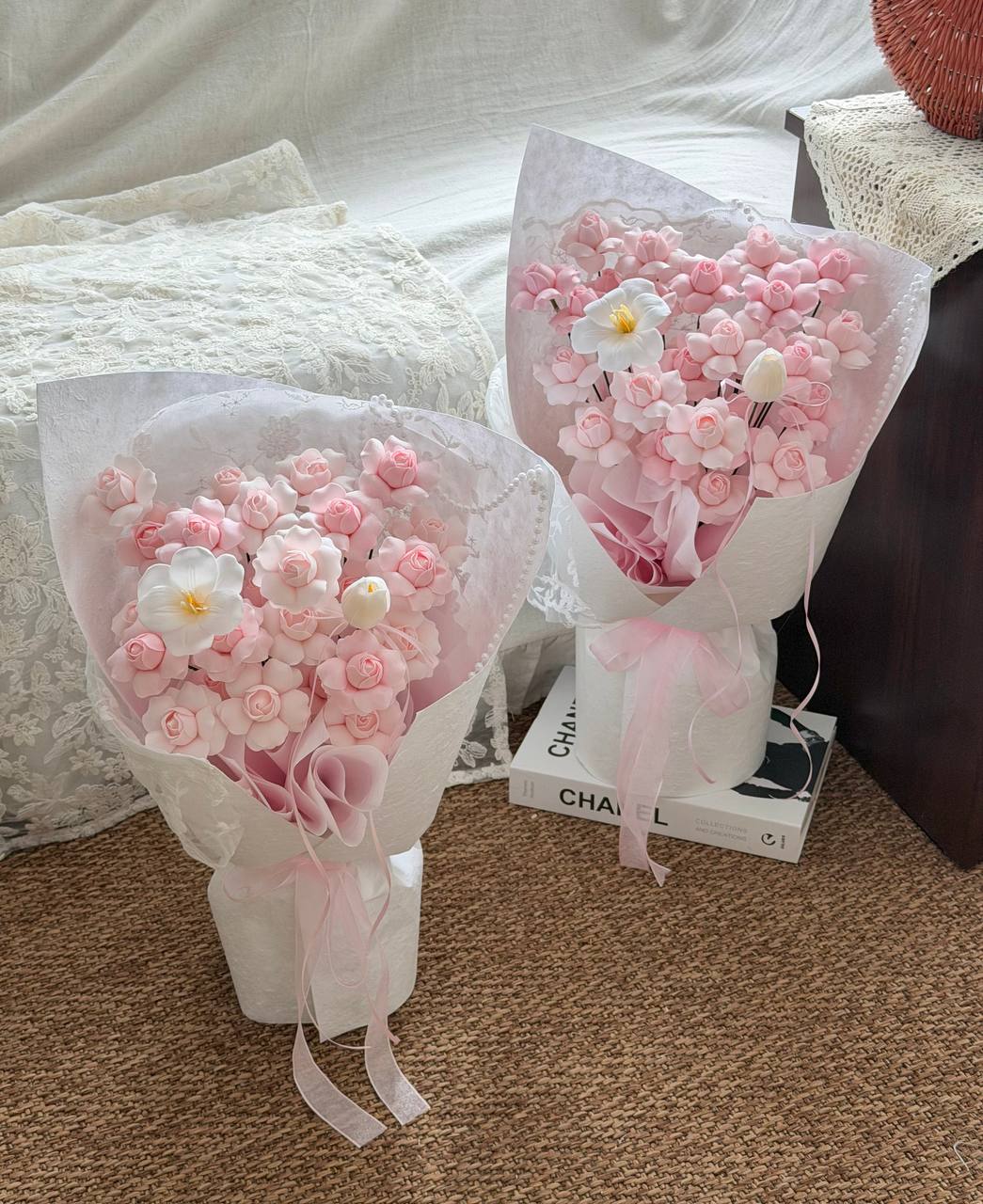 Two wrapped flower bouquets made of pink and white soap flowers kiss of fairy rose spray, wrapped in sheer white paper with pink ribbons, placed on a carpeted floor in a bedroom setting.
