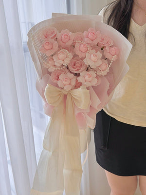 A bouquet of pink kiss of fairy rose spray wrapped in sheer blush and cream paper with a large cream ribbon bow, held by a person wearing a cream top and black skirt in front of white curtains.