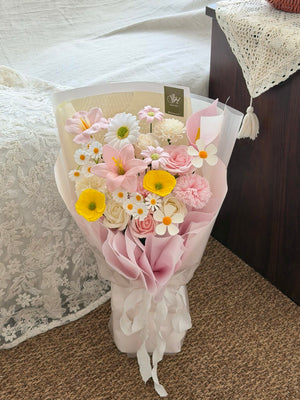 A wrapped bouquet soap flower of pink, white, and yellow flowers in sheer pink paper wrapping, tied with a white ribbon, placed on a carpeted floor next to a bed.