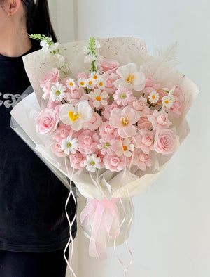 A bouquet of pink and white flowers wrapped in cream paper and tied with a pink ribbon, held by a person wearing a black shirt against a plain light background.