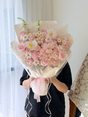 A person in a black shirt holds a large bouquet of kiss of fairy rose spray soap flower wrapped in cream paper with a pink ribbon, standing indoors near a window with sheer curtains.