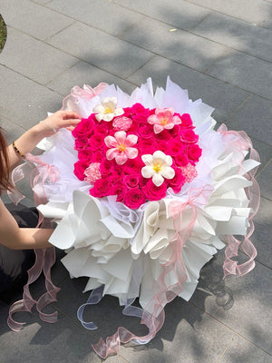 A large heart-shaped flower bouquet made of bright hot pink roses with white tulip and carnation, wrapped in white ballet wrapper and pink sheer ribbons, being held by a person outdoors on a gray paved surface.