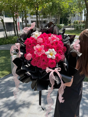 A large, vibrant flower bouquet held by a person outdoors. The bouquet features hot pink roses, tulips and carnation, wrapped in black paper with light pink ribbons tied in bows.