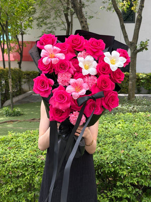 A person outdoors holding a large bouquet of hot pink roses, dyed tulip, white tulip and dyed carnation wrapped in black paper with black ribbons. The background shows green shrubs and trees.
