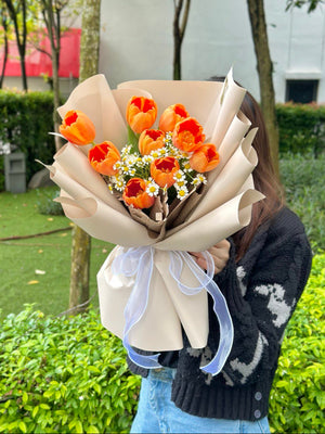 In Kuala Lumpur, a woman wearing a black cardigan holds a vibrant bouquet of orange tulips for her friend's birthday.