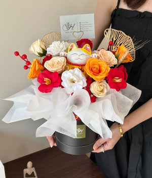 A festive floral arrangement featuring red, white, and orange soap roses with a lucky cat figurine in the center, perfect for prosperity and celebration.