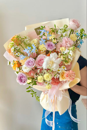 A large, dreamy bouquet filled with pastel roses, carnations, daisies, and blue delphiniums, perfect for celebrations or heartfelt gifts.