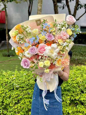 A lush, garden-style bouquet with layered pastel blooms and delicate wrapping, held by a woman in a navy top and denim skirt.