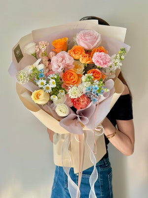 A vibrant mixed flower bouquet featuring orange roses, pale pink carnations, chamomile, and blue delphiniums, wrapped in pastel paper.