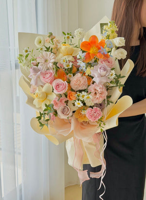 A person holding a large, vibrant floral bouquet with pink, orange, yellow, and white flowers wrapped in cream paper with a pink ribbon, set against a bright, softly lit window background.