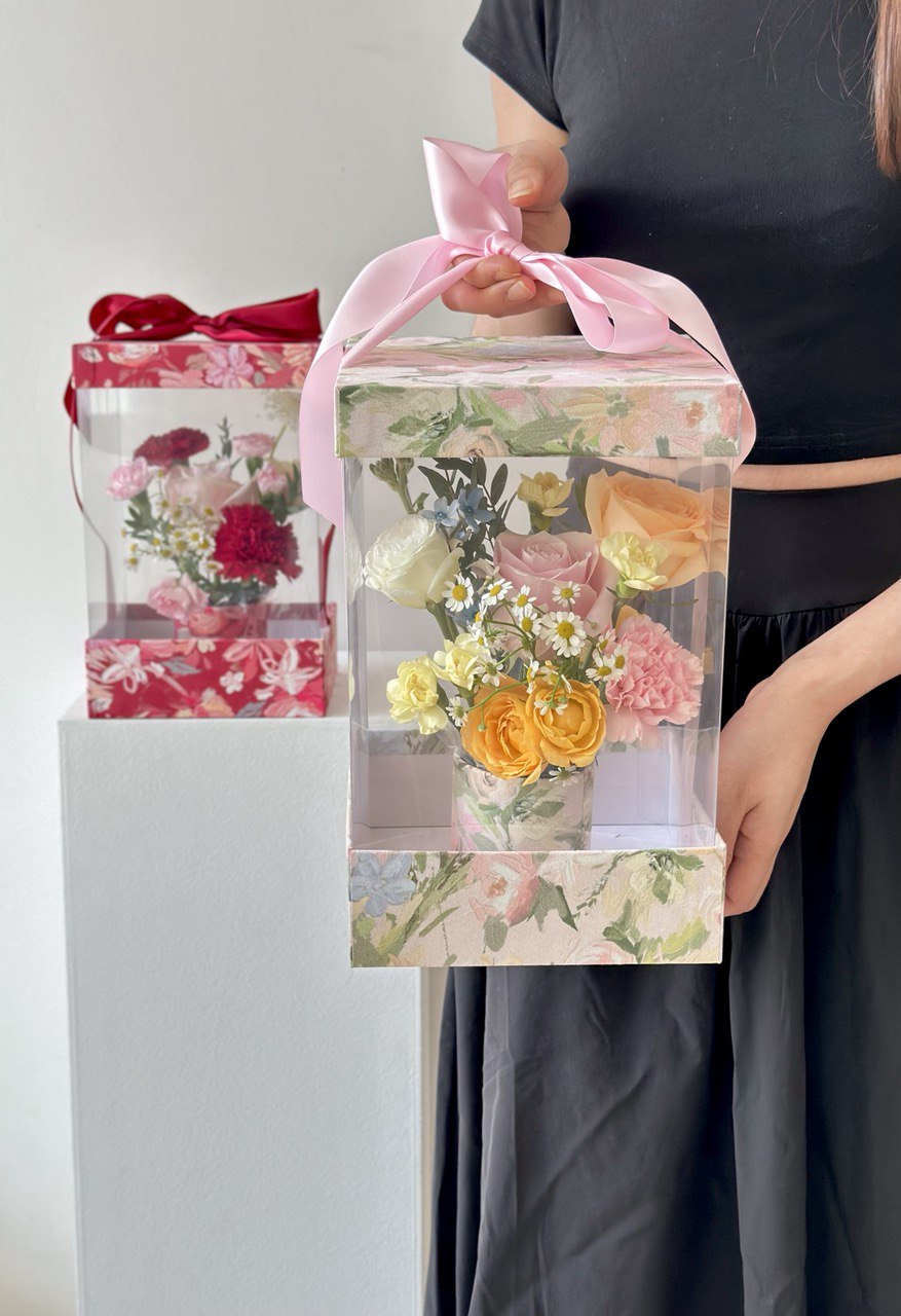 A young woman in a black dress presents a box of flowers and a Mother's Day card, celebrating her mother on Mother's Day 2025.