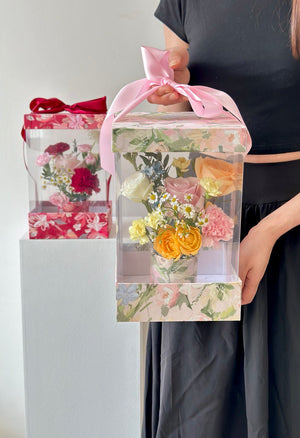 A young woman in a black dress presents a box of flowers and a Mother's Day card, celebrating her mother on Mother's Day 2025.