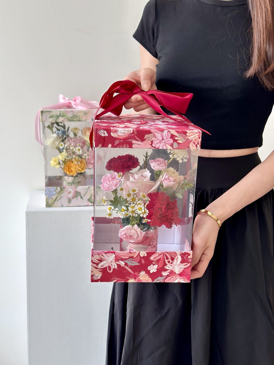 A girl wearing a black dress holds a floral gift box and a Mother's Day card, ready to honor her mother on this special day.
