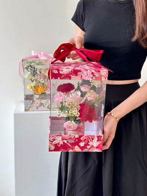 A girl wearing a black dress holds a floral gift box and a Mother's Day card, ready to honor her mother on this special day.