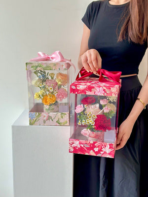 A woman in a black dress carries a box filled with flowers and a Mother's Day card, expressing love for her mother in 2025.