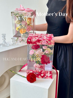 A girl in a black dress holds a box of flowers and a Mother's Day card, smiling as she prepares to surprise her mother.