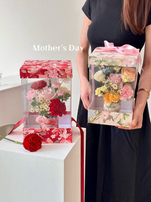 A girl in a black dress holds a box of flowers and a Mother's Day card, joyfully preparing to celebrate her mother.