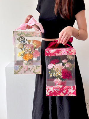 A girl in a black dress holds a box of flowers and a Mother's Day card, joyfully preparing to celebrate her mother.