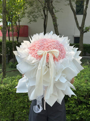 A large bouquet of 52 pink roses wrapped in white paper with a long white ribbon bow, held outdoors in front of green hedges and trees.