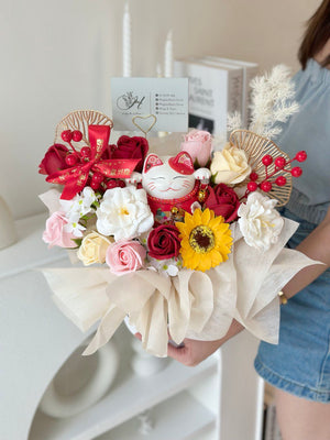 A girl wearing a blue top is holding a white box with a floral bouquet and a lucky cat on top, marking a business opening event in Kuala Lumpur.