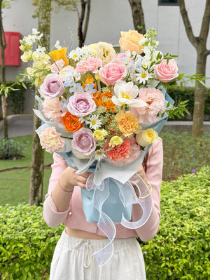 Pastel rose and carnation bouquet with mixed spring flowers wrapped in light blue paper.