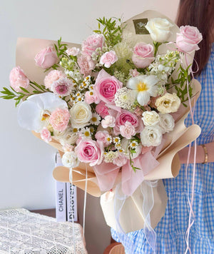 A large pastel bouquet featuring pink roses, carnations, and white orchids, held in front of a person wearing a blue gingham dress.