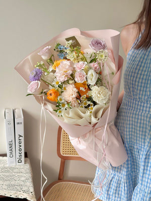 A vibrant and romantic floral arrangement held by a woman in a blue gingham dress, showcasing a mix of blush, lilac, and orange blooms.