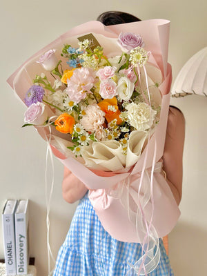 A pastel-toned bouquet featuring roses, ranunculus, and chamomile, elegantly wrapped in soft pink paper with flowing ribbons.