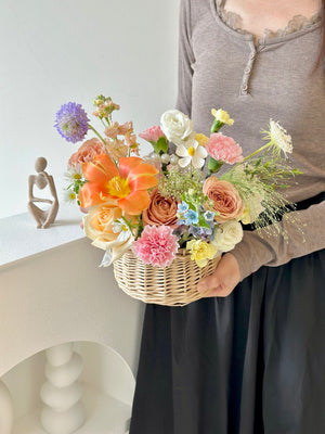 A woman in a brown top carries a basket of flowers, with an additional basket on the table, ideal for various occasions.