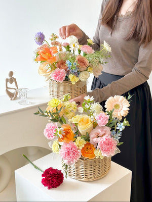 A woman in a brown top holds a basket of flowers, with another basket placed on a table, perfect for any occasion.