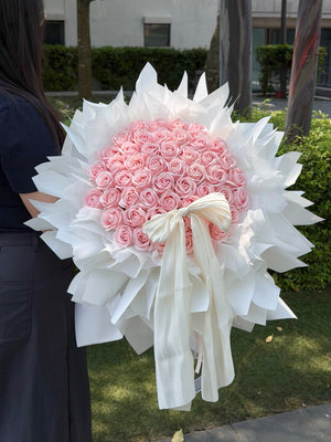 A large round-shaped bouquet of 52 pink roses soap flower wrapped in white paper and tied with a white ribbon, held by a person standing outdoors on grass with trees and a building in the background, perfect for proposal gift.