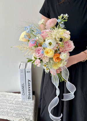 Hand-tied bridal bouquet featuring yellow, pink, orange, blue, and white blooms with lush greenery.