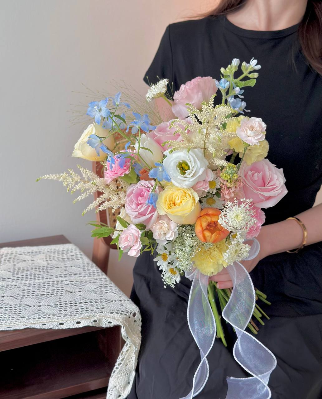 Woman in black dress holding a colorful bridal bouquet with pink roses, buttercup rose, carnations, astilbe and delicate fillers tied with a white ribbon.