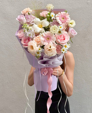 A person holding a large, beautifully arranged flower bouquet with pink roses, white lilies, and daisies wrapped in light purple paper with pink ribbons, set against a neutral gray wall.