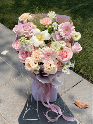 A elegant bouquet of pink, peach, white, and light purple flowers wrapped in a matching light purple paper sleeve with a long pink ribbon, placed on a gray stone path with green grass in the background.