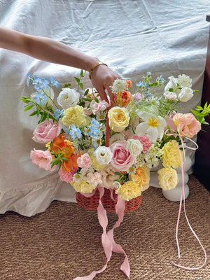 Bouquet of flowers in a basket with a hand reaching towards it, on a white sheet background.