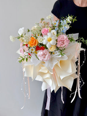 A woman in a black dress holding a large pastel bouquet with roses, tulips, carnations, and delicate filler flowers wrapped in cream-colored paper.