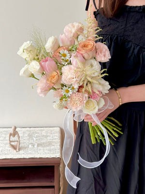 Bride holding a romantic blush-toned bridal bouquet with roses, tulips, carnations, and delicate white fillers tied with a sheer ribbon.