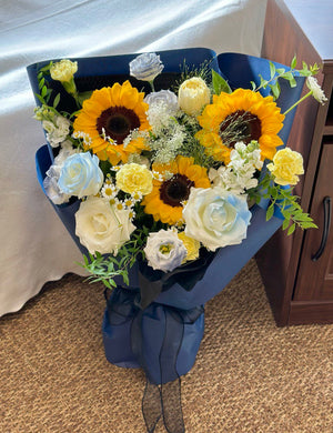Bouquet of sunflowers, white roses, and greenery in a blue wrap on a carpeted floor, perfect for graduation in Malaysia.