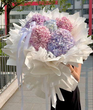 A large bouquet of pastel hydrangea flowers wrapped in white ballet style paper with white ribbons, held by a person wearing a black dress outdoors on a paved walkway with trees and buildings in the background.
