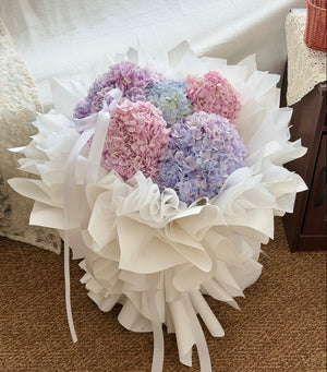 A bouquet of Large hydrangea flowers in pastel pink, lavender, and light blue, wrapped in white ballet style wrapper with long white ribbons, placed on a brown carpeted floor in an indoor setting.