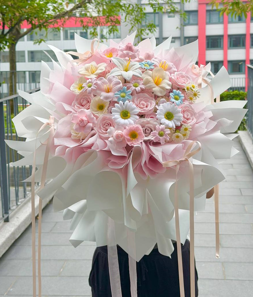 A large, elaborate bouquet of pink and white flowers with small pastel blue and yellow accents, wrapped in white paper and held by a person wearing black pants on an outdoor walkway.