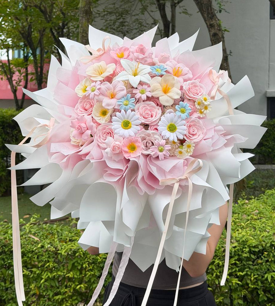 A large, vibrant flower bouquet wrapped in white paper with pink roses, daisies, and small blue and yellow flowers, held by a person outdoors in front of green hedges and trees.