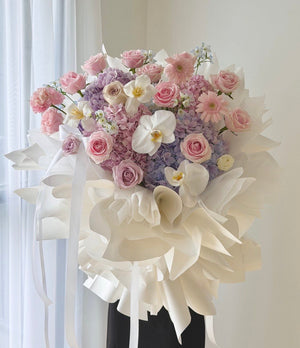 A large, lush floral arrangement featuring pink and purple roses, white orchids, and hydrangeas, wrapped in elegant white paper and tied with long white ribbons, displayed on a black stand against a soft, light-colored background.