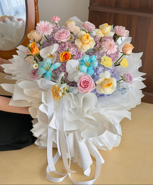 A large, vibrant bouquet of multicolored flowers and hydrangea wrapped in white paper with long white ribbons, held by a person whose arm and hand are partially visible on the left, set against an indoor background.