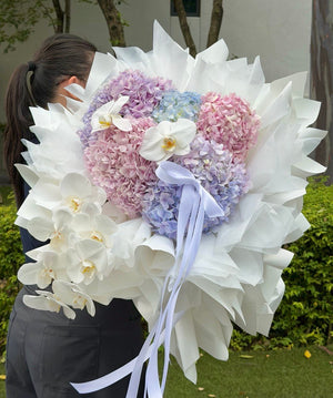 A large heart-shaped bouquet of pastel hydrangeas in pink, purple, and blue, wrapped in white wrapper paper with white orchid flowers and a light blue ribbon, held by a person outdoors.