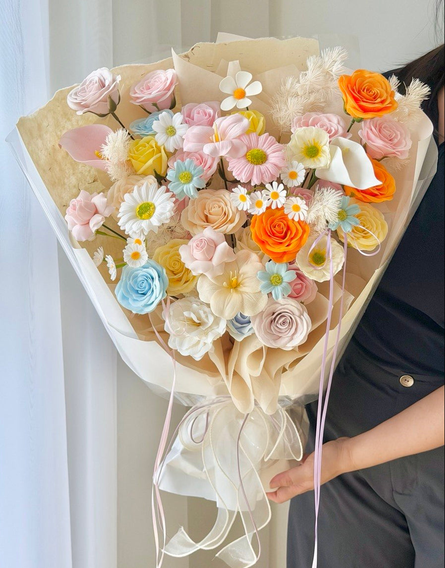 A large, beautifully arranged floral bouquet held by a person wearing a black top. The bouquet features a mix of pastel pink, orange, yellow, and light blue artificial flowers, white daisies, and dried fluffy accents, wrapped in beige paper with a sheer cream ribbon.