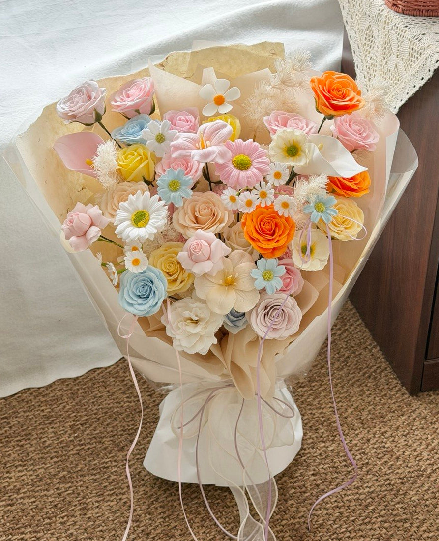 A pastel-colored artificial flower bouquet wrapped in beige paper, featuring roses, daisies, and small blossoms in pink, orange, yellow, blue, and white, placed on a carpeted floor next to a white draped surface.