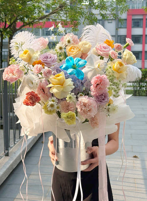 A person outdoors holding a large, vibrant bouquet of multicolored flowers, including pink, yellow, peach, lavender, and blue tulip, placed in a silver flower box with feather accents and ribbons, in a silver bucket vase.
