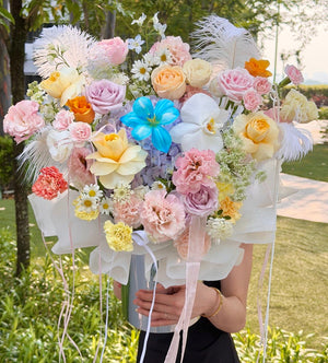 A large, vibrant flower bouquet held by a person in a black dress outdoors. The bouquet features a mix of pastel pink, yellow, orange, and white flowers, including roses, carnations, hydrangeas, and white feathers, wrapped in white tissue paper with long pink ribbons.